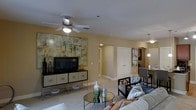This single-story residence in Irving offers a thoughtfully designed open-concept layout that connects the living, dining, and adjacent areas in a cohesive and functional arrangement. The living area serves as the focal point of the space, anchored by a ceiling fan with integrated lighting that illuminates the room with a warm, comfortable glow. Soft, neutral-toned carpeting extends throughout, complementing the warm beige walls and contributing to the overall inviting ambiance.

A media console with geometric drawer fronts serves as the entertainment centerpiece, topped with a flat-screen television and flanked by decorative accents. Above it, a large-scale abstract artwork introduces rich gold and green tones, adding visual depth and a contemporary artistic touch. The sitting area features a plush sofa adorned with patterned accent pillows, alongside a sleek glass-topped coffee table styled with decorative vases.

Adjacent to the living space, a dining area is illuminated by pendant lighting, with a bistro-style table and chairs offering an intimate setting. Hallways lead to additional rooms, including what appears to be a closet or storage area, reinforcing the space's practical and well-organized design.