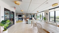This single-floor space is a thoughtfully designed open-plan lounge that combines eclectic bohemian aesthetics with a bright, welcoming atmosphere. Light-washed hardwood flooring runs throughout, complementing the white tile-panel ceiling fitted with warm gold woven pendant lights that add a tropical, resort-inspired character.

The layout is organized into several distinct seating zones that flow seamlessly into one another. A communal dining area features a large natural wood table surrounded by white rattan chairs, positioned near floor-to-ceiling windows that frame expansive outdoor views and flood the interior with natural light. A relaxed lounge area showcases wicker peacock chairs, rattan ottomans, leather pouf seating, and a soft mint sectional sofa, all accented by lush tropical plants that reinforce the space's organic, laid-back energy.

A cozy nook with terracotta-tiled banquette seating in deep red upholstery provides an intimate dining or gathering spot, while a separate casual lounge corner features a teal sofa, bold orange accent chair, and colorful wall art, adding a playful, energetic contrast. Open black metal shelving units display curated décor and greenery, serving as stylish room dividers. White brick-textured walls along the window-lined perimeter further enhance the airy, coastal-inspired ambiance throughout.