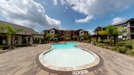 This outdoor pool area offers a well-appointed retreat designed for relaxation and leisure. At its centerpiece is a generously sized freeform pool with crystal-clear water, bordered by an expansive paver deck finished in neutral stone tones that provide both durability and visual appeal. The deck is furnished with lounge chairs and seating areas shaded by vibrant blue umbrellas, creating comfortable spots to unwind under open skies. A covered lounge pavilion anchors one side of the space, featuring floor-to-ceiling glass panels, ceiling fans for airflow, and wicker seating arranged on a tile floor — offering a sheltered yet open-air environment with direct views of the pool. Natural stone columns with decorative wall-mounted lanterns frame the transition between the covered area and the pool deck, adding a warm architectural character. Lush tropical plantings, including tall palms and ornamental grasses in colorful accent pots, are thoughtfully placed throughout, contributing to the resort-like ambiance. A wrought iron perimeter fence encloses the space, ensuring privacy and security. The surrounding multi-story buildings with stone and stucco facades complete the setting, providing an attractive and cohesive backdrop to this vibrant outdoor amenity.