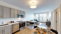 This thoughtfully designed single-floor residence offers a seamless blend of modern aesthetics and practical living. The open-concept layout connects the kitchen, dining, and living areas into one cohesive space, ideal for both everyday living and entertaining. The kitchen is outfitted with grey shaker-style cabinetry, a classic white subway tile backsplash, and a full suite of stainless steel appliances, including a range, microwave, dishwasher, and refrigerator. Light wood-tone flooring flows throughout the space, contributing to a warm and airy atmosphere. The dining area accommodates a round table with seating for multiple guests, positioned conveniently between the kitchen and living room. The living area benefits from large windows dressed with horizontal blinds, allowing natural light to fill the room while maintaining privacy. The bedroom is a serene retreat, featuring generous window coverage, light-toned walls, and ample space. The bathroom presents a clean, contemporary design with a floating wood-grain vanity, an undermount sink, a large mirror, and a tiled shower-tub combination. Throughout the unit, the neutral color palette and modern finishes create a cohesive, inviting environment.