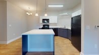 Located at 4830 Wescott Blvd in Summerville, South Carolina, this single-floor unit offers a thoughtfully designed layout with a clean, modern aesthetic throughout. The kitchen is a standout feature, showcasing a two-tone design with crisp white upper cabinetry and bold navy-blue lower cabinets, paired with light countertops and a full suite of black appliances including a range, microwave, dishwasher, and refrigerator. A central island with a white surface and matching blue base adds additional workspace and visual interest, while pendant and chandelier lighting overhead creates an inviting atmosphere. Light wood-style flooring flows through the kitchen and into the open living area, which benefits from natural light through windows with blinds, a ceiling fan, and a built-in shelving nook ideal for display or storage. The bedroom features plush carpeting and a window overlooking greenery outside. The primary bathroom includes a white vanity with sink, a tub-shower combination, and hardwood-style flooring, with access to a walk-in closet featuring wire shelving. A dedicated laundry room provides washer and dryer hookups, additional overhead shelving, and a pet door, rounding out this well-appointed and functional space.