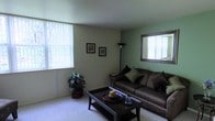 This single-story space centers around a comfortable living room that combines functional design with a relaxed aesthetic. The room is finished with wall-to-wall carpeting underfoot and features two-tone walls, with soft green accent walls complementing lighter neutral tones throughout. Natural light is a notable feature, with vertical blinds adorning two windows — one large floor-length window and a second framed by a decorative mirror — allowing daylight to filter warmly into the space. The framed mirror on the green accent wall adds depth to the room, creating a sense of expanded space. Decorative framed artwork is displayed on the walls, adding a personal touch to the environment. The living area accommodates a full-sized sofa dressed with accent pillows, paired with a dark wood coffee table complete with a lower storage shelf. A tall arc floor lamp provides additional ambient lighting, while a potted leafy plant in the corner introduces a natural, organic element. The overall atmosphere is cozy and inviting, with a clean, modest design that prioritizes comfort and practicality.