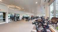 Located at 3955 Design Center Dr in Palm Beach Gardens, FL, this professionally appointed fitness center offers a generous, single-floor exercise space designed to support a comprehensive workout experience. The facility features a bright, open layout with soaring ceilings and an abundance of natural light streaming through large floor-to-ceiling windows that frame views of a resort-style pool area surrounded by palm trees, creating an energizing and inviting atmosphere.

The exercise room is equipped with an extensive array of cardio machines, including Peloton cycling bikes, treadmills, ellipticals, and stair climbers, complemented by a full selection of strength-training equipment such as cable machines, selectorized weight machines, and adjustable benches. A dedicated free weights area offers a complete dumbbell rack alongside medicine balls, kettlebells, and stability balls for functional and dynamic training.

The interior design features a clean, modern aesthetic with a neutral color palette of white walls and light-toned flooring. Geometric hexagonal pendant lighting fixtures add a sophisticated architectural touch overhead. Full-length mirrors along the walls enhance the sense of space and assist with form during workouts. A motivational wall graphic reading "Be Strong. Be Tough. Be Daring. Be You." reinforces the facility's energetic character. A water fountain is also conveniently accessible within the space.