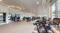 Located at 3955 Design Center Dr in Palm Beach Gardens, FL, this professionally appointed fitness center offers a generous, single-floor exercise space designed to support a comprehensive workout experience. The facility features a bright, open layout with soaring ceilings and an abundance of natural light streaming through large floor-to-ceiling windows that frame views of a resort-style pool area surrounded by palm trees, creating an energizing and inviting atmosphere.

The exercise room is equipped with an extensive array of cardio machines, including Peloton cycling bikes, treadmills, ellipticals, and stair climbers, complemented by a full selection of strength-training equipment such as cable machines, selectorized weight machines, and adjustable benches. A dedicated free weights area offers a complete dumbbell rack alongside medicine balls, kettlebells, and stability balls for functional and dynamic training.

The interior design features a clean, modern aesthetic with a neutral color palette of white walls and light-toned flooring. Geometric hexagonal pendant lighting fixtures add a sophisticated architectural touch overhead. Full-length mirrors along the walls enhance the sense of space and assist with form during workouts. A motivational wall graphic reading "Be Strong. Be Tough. Be Daring. Be You." reinforces the facility's energetic character. A water fountain is also conveniently accessible within the space.