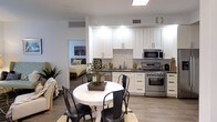 This single-floor space showcases a well-designed open-concept layout that integrates the kitchen and dining area into a cohesive living environment. The kitchen is outfitted with crisp white shaker-style cabinetry that extends to the ceiling, providing generous storage throughout. Dark countertops create a striking contrast against the white cabinetry, while a light-toned tile backsplash adds texture and visual interest. Stainless steel appliances, including a gas range, over-the-range microwave, and dishwasher, contribute a sleek, modern aesthetic. Recessed lighting beneath the upper cabinets ensures the workspace is well illuminated. The dining area sits adjacent to the kitchen and features a round white table paired with black metal chairs, grounded by a woven area rug atop wood-look flooring that runs throughout the space. A bonsai tree centerpiece adds a natural, decorative touch. An open doorway connects this area directly to the bedroom, which is visible in the background, enhancing the sense of flow and connectivity. The overall ambiance is clean, modern, and efficiently organized.