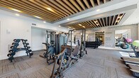 This fitness center offers a thoughtfully designed workout environment that balances aesthetic appeal with comprehensive functionality. The space is defined by a striking slatted wood ceiling detail with recessed lighting that runs throughout, adding warmth and architectural character to the interior. Patterned carpet tiles in a neutral herringbone-style design cover the floors, providing comfort and visual texture underfoot.

The strength training area is well-equipped with a full lineup of cable machines, multi-functional weight stations, adjustable benches, and an extensive dumbbell rack ranging across multiple weight increments. A large mirrored wall expands the sense of space and is accented by a bold typographic mural featuring fitness-related words, reinforcing the motivational atmosphere.

The cardio zone is positioned near floor-to-ceiling windows that invite abundant natural light and outdoor views, housing a row of stair climbers, treadmills, and elliptical machines. A textured stacked-stone accent wall adds an earthy, spa-like contrast to the modern design. Yoga and stability balls, a beverage station, and additional free weight storage round out the amenities. The overall ambiance is modern, energizing, and well-organized.