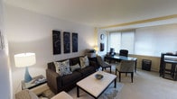 This single-floor space offers a well-balanced combination of relaxation and productivity in a thoughtfully arranged layout. The main living area is anchored by a plush dark brown sofa dressed with decorative patterned cushions, positioned across from a sleek white rectangular coffee table that grounds the seating arrangement. A textured area rug defines the lounge zone and adds warmth underfoot.

A trio of black botanical wall art panels creates a striking focal point on one wall, lending a modern aesthetic to the interior. The transition between the lounge and workspace is seamless, with the office area featuring a glass-topped desk, an ergonomic office chair, and a side chair — ideal for meetings or focused work. A multi-function printer and organized desk accessories suggest a fully equipped workspace.

Natural light filters through a large window fitted with horizontal blinds, brightening the neutral-toned walls and light carpet throughout. Warm accent lighting from a table lamp adds a soft glow to complement the overhead illumination. The space features a clean architectural finish with a subtle raised ceiling detail that adds visual interest. Overall, the interior exudes a contemporary, professional, and inviting atmosphere suited for both work and leisure.