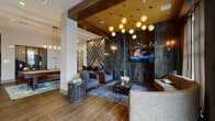 This sophisticated single-floor amenity space makes a striking first impression with its thoughtfully layered design and upscale finishes. The central lounge area is anchored by a dramatic floor-to-ceiling dark stone tile feature wall, upon which a flat-screen TV is mounted and a cascading cluster of amber globe pendant lights descends from a warm gold-toned ceiling recess framed by exposed dark wood beams. Wood-look tile flooring runs throughout, grounding the space with a warm, earthy tone. Plush seating arrangements, including modern upholstered chairs and a deep sofa, are complemented by artisan side tables and richly patterned area rugs.

An eye-catching abstract stone wall art installation adds a bold focal point, while a dedicated work and seating nook nearby provides a quieter retreat. The games area features a full-sized pool table set atop a vivid blue marbled rug beneath a sleek rectangular pendant fixture, with access to a private room beyond glass-paneled doors.

The kitchen and bar area showcases an expansive granite island with built-in sink and bar seating, illuminated by sculptural gold cage pendants. A dimensional geometric tile backsplash and large-format hexagonal floor tile elevate the space. Adjacent, a cozy dining nook benefits from floor-to-ceiling windows, rich wood panel walls, living moss wall art, and tufted banquette seating, creating an inviting atmosphere filled with natural light.