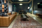 Located at 1143 First Ave S in Birmingham, Alabama, this single-floor space masterfully blends raw industrial architecture with sophisticated interior design. The building retains its original structural character through exposed concrete columns, an open ceiling with visible ductwork and track lighting, and polished concrete floors — all hallmarks of a thoughtfully converted commercial or warehouse property.

The main lounge area serves as the centerpiece of the space, anchored by a rich cognac leather sofa and complementary dark velvet seating arranged around a wood-and-glass coffee table atop a layered vintage area rug. Deep navy and dark teal walls lend a moody, upscale ambiance, while globe pendant lights and strategically placed floor lamps provide warm, inviting illumination.

A dramatic floor-to-ceiling modular shelving wall — constructed of steel and warm wood panels — displays an eclectic curation of decorative objects, adding both visual depth and artistic character. A wall-mounted flat-screen TV with a mid-century media console anchors an adjacent seating vignette.

The back of the space opens into a more expansive raw area, suggesting additional functional potential. Heavy-duty industrial doors with viewing windows are also present, hinting at utility or storage access. The overall atmosphere is bold, curated, and distinctly urban.
