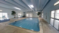 This single-level indoor pool facility offers a welcoming aquatic environment designed for year-round use. The centerpiece is a generously sized rectangular swimming pool with clear blue water, accessible via stainless steel handrails. Depth markings are visible along the pool's edge, indicating a shallow end suitable for a range of swimmers. Adjacent to the main pool sits a hot tub, providing a relaxing complement to the swim area.

The enclosure features a light blue ceiling with exposed beams and multiple skylights that flood the space with natural light, creating an airy, open atmosphere. Large windows along the perimeter walls further enhance the brightness while offering views to the surrounding exterior. The pale blue walls are decorated with painted tropical palm trees, adding a resort-like ambiance to the indoor setting.

The pool deck is finished in neutral-toned concrete, providing a practical and slip-resistant surround. A small seating area with chairs is positioned near the hot tub, offering a comfortable spot to relax. The overall design balances functionality with a cheerful, leisure-focused aesthetic, making it a versatile facility for both recreational swimming and relaxation.