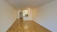 Located at 389 South End in Battery Park City, New York, this single-floor residence offers a clean, open layout designed for straightforward, functional living. The main living area is spacious and unobstructed, finished with light-toned hardwood flooring that runs throughout, lending warmth and visual continuity to the space. Neutral white walls and flush-mount ceiling lighting keep the interior feeling bright and airy.

The kitchen is positioned at the rear of the open-plan living area, partially separated by a half-wall peninsula that doubles as a natural divider without fully enclosing the space. The kitchen features white cabinetry, a stainless steel refrigerator, and pendant lighting suspended from above, providing both task illumination and a subtle decorative touch. A stainless steel sink and countertop workspace complete the functional cooking area.

An entryway with a full-height door is visible adjacent to the kitchen, providing access to additional areas of the unit. The overall ambiance is minimalist and neutral, offering a blank canvas that adapts well to a variety of interior styles. The straightforward design and consistent finishes give the space a cohesive, move-in-ready character.