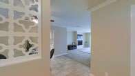 This single-story residence offers a welcoming and functional layout designed for comfortable everyday living. The entryway features neutral-toned tile flooring that transitions seamlessly into the carpeted living area, creating a clear visual distinction between spaces while maintaining a cohesive aesthetic throughout.

The living room serves as the heart of the home, anchored by a classic fireplace framed by a clean, traditional surround. Crown molding adds a refined architectural detail along the ceiling line, lending a polished finish to the room. The walls are finished in a warm, neutral tone that complements the light-colored carpet and contributes to the bright, airy atmosphere of the space.

A media console is positioned in front of the fireplace, providing a practical and centrally located entertainment area. Natural light filters in through a window visible near the far wall, enhancing the open and inviting feel of the living room.

Adjacent to the entryway, a glimpse into a connected room — likely a dining area or kitchen — hints at a well-connected floor plan that supports easy flow between living spaces. Decorative elements near the entry corridor add a touch of personality to the transition area. Overall, this home presents a clean, move-in ready interior with classic detailing throughout.
