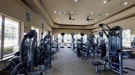 This fitness center offers a comprehensive workout environment designed to accommodate a variety of exercise needs. The single-floor layout is thoughtfully organized, with cardio equipment — including treadmills, ellipticals, and stair climbers — arranged toward the rear of the space, while a robust selection of strength training and resistance machines lines both sides of the room. The flooring is finished with durable carpet, providing cushioning underfoot throughout the workout area. Elevated ceilings with a tray detail lend an open, spacious feel to the interior, while ceiling fans and recessed lighting ensure a comfortable and well-lit atmosphere. Large windows along the back wall invite abundant natural light and offer views to the outdoors, creating an energizing environment. Wall-mounted televisions are positioned for visibility from the cardio equipment, adding entertainment options during workouts. Motivational posters and wall clocks are strategically placed throughout the space. The overall ambiance is clean, functional, and inviting, making this fitness center a well-appointed facility suited for a full range of daily exercise routines.