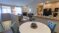 This two-story residence is designed around an open-concept main living area that seamlessly connects the dining, living, and kitchen spaces. The living room serves as a central gathering point, anchored by a fireplace with an arched architectural surround and framed by a large window dressed in sheer white curtains that invite natural light into the space. Warm hardwood flooring extends throughout, lending continuity and a sense of warmth across the open floor plan.

The dining area features a round white table with tufted upholstered chairs in a muted teal tone, positioned to overlook both the living room and the kitchen beyond. The kitchen is set slightly elevated behind a granite-topped breakfast bar, offering a functional pass-through between cooking and entertaining areas. Light wood cabinetry, granite countertops, and black appliances complete the kitchen's practical yet polished aesthetic.

Vaulted ceilings rise above portions of the main level, enhancing the sense of openness and volume throughout. The neutral warm-gray wall palette throughout creates a cohesive, calm ambiance, while recessed lighting supplements the natural light. The second floor adds additional living space to this well-appointed, thoughtfully designed home.