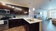 This single-floor residence showcases a thoughtfully designed open-concept layout, where the kitchen flows seamlessly into the living and sleeping areas. The kitchen is a standout feature, outfitted with rich dark espresso cabinetry, light quartz-style countertops, and a subway tile backsplash. A peninsula with a stainless steel undermount sink and elegant pendant lighting serves as both a functional workspace and a casual dining area, complete with bar stool seating. Stainless steel appliances, including a range and microwave, add a polished, contemporary touch. Hardwood-style flooring runs throughout the unit, lending warmth and visual continuity to the space. The open layout provides a clear sightline from the kitchen into the living area and bedroom, creating an airy, connected feel. The bedroom area is comfortably proportioned and positioned near a walk-in closet, while a dedicated nook with a built-in desk offers a practical workspace. The bathroom features a walk-in shower with a curtain enclosure and coordinating hardwood-style flooring. Track lighting and pendant fixtures provide layered illumination throughout, enhancing the modern, functional character of this well-appointed residence.