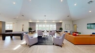 This well-appointed single-floor amenity space offers a generous, open layout designed for both relaxation and productivity. The main living area is anchored by soaring vaulted ceilings and light-toned herringbone-patterned flooring that flows throughout, creating a cohesive and airy atmosphere. A bold orange sectional sofa faces a wall-mounted television, while a separate lounge cluster of lavender-gray armchairs arranged around a glass coffee table provides additional seating. Natural light pours in through large windows dressed with sheer roller shades and floor-length draping curtains, offering views of the surrounding outdoor space. An arched window accent adds a distinctive architectural touch to one end of the living area.

The dining area features multiple dining tables positioned along a bank of oversized windows, ideal for casual gatherings. Adjacent to this is the kitchen, which showcases a striking dark marble-top bar counter with live-edge plant accents and sleek wood bar stools, illuminated by globe pendant lights with brass fixtures. Stainless steel appliances, white tile backsplash, and a beverage station complete the kitchen's modern aesthetic.

The dedicated office is compact yet functional, featuring a built-in dual-workstation desk equipped with monitors and a printer, lit by modern pendant lamps against a patterned accent wall.