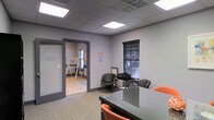 This single-story commercial office space offers a functional and professional environment suited for business operations. The main area features a sleek glass conference table surrounded by orange and black chairs, creating a modern focal point ideal for meetings and collaborative work. The room is finished with neutral gray walls and carpeted flooring, contributing to a clean, professional aesthetic.

A drop ceiling with recessed fluorescent panel lighting provides consistent, even illumination throughout the space. A window fitted with dark horizontal blinds allows for natural light control while maintaining privacy. The open doorway connects to an adjacent room, visible in the background, which features hard flooring, a whiteboard or easel, and additional natural light from a window — suggesting use as a training room, breakout space, or secondary office.

A frosted glass panel beside the interior door adds a modern design touch while allowing light to pass between areas. A printer station and additional seating are positioned along one wall, enhancing the room's utility. Overall, the space balances practicality with a contemporary office design, making it well-suited for professional or small business use.