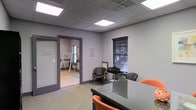 This single-story commercial office space offers a functional and professional environment suited for business operations. The main area features a sleek glass conference table surrounded by orange and black chairs, creating a modern focal point ideal for meetings and collaborative work. The room is finished with neutral gray walls and carpeted flooring, contributing to a clean, professional aesthetic.

A drop ceiling with recessed fluorescent panel lighting provides consistent, even illumination throughout the space. A window fitted with dark horizontal blinds allows for natural light control while maintaining privacy. The open doorway connects to an adjacent room, visible in the background, which features hard flooring, a whiteboard or easel, and additional natural light from a window — suggesting use as a training room, breakout space, or secondary office.

A frosted glass panel beside the interior door adds a modern design touch while allowing light to pass between areas. A printer station and additional seating are positioned along one wall, enhancing the room's utility. Overall, the space balances practicality with a contemporary office design, making it well-suited for professional or small business use.