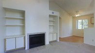 This single-story space is defined by its open, airy living area highlighted by vaulted ceilings that lend a sense of generous vertical volume. The focal point of the main living space is a classic tile-surround fireplace framed by a bold black insert, flanked on both sides by built-in shelving and cabinetry. The shelving units offer practical open display storage above and closed cabinet storage below, seamlessly integrated into the wall design on either side of the fireplace. The walls are finished in a clean, neutral white tone that reflects light and enhances the brightness of the interior. The flooring transitions between light tile near the fireplace hearth and carpet in the main living zone, providing both durability and comfort underfoot. A HVAC vent is visible near the upper wall, indicating a central climate control system. The space flows openly into an adjoining area, suggesting a connected, functional layout. The overall ambiance is calm and understated, offering a neutral canvas well-suited for a variety of interior styles.