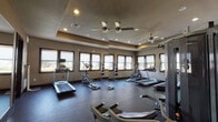 This fitness center offers a comprehensive workout environment designed for functionality and comfort. The space is accessed through a dark-stained wood-and-glass entry door, lending a polished first impression. Inside, the main workout area is finished with durable dark rubber flooring, neutral-toned walls, and a tray ceiling accented by recessed lighting and dual ceiling fans that ensure proper air circulation throughout.

Cardio equipment is prominently featured, including multiple treadmills and elliptical cross-trainers positioned along the perimeter to take advantage of the natural light streaming in through a generous row of windows fitted with roller shades. A dedicated strength-training station anchors one area of the room, complemented by a medicine ball rack for functional fitness exercises. A rowing machine and free weights, including dumbbells, round out the available equipment for a full-body training experience.

Full-length mirrors line one wall, visually expanding the space and allowing users to monitor their form during workouts. The abundant window coverage provides views to the outdoors and keeps the atmosphere bright and energizing. The overall layout is open and thoughtfully organized, offering ample floor space for movement and stretching, making this fitness center a well-appointed facility suited for a variety of training routines.