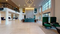 Located at 2395 Herodian Way SE in Atlanta, Georgia, this expansive single-floor amenity space delivers a polished, resort-inspired atmosphere designed for both socializing and relaxation. The centerpiece of the space is a dramatic lobby featuring soaring double-height ceilings, large-format light stone tile flooring, and a striking reception desk finished in teal and warm wood tones. A floor-to-ceiling textured wall panel serves as an architectural focal point behind the desk, flanked by decorative geometric accents. An eye-catching cascading chandelier adds warmth and visual drama overhead, while an upper mezzanine with black iron railings overlooks the main floor below.

Adjacent lounge areas offer varied seating arrangements, including tufted white modular chairs, bold navy wingback chairs, and intimate clusters of emerald green high-back seating positioned near large steel-framed windows. A media lounge features a large-format television set against vibrant teal wallcovering. A communal gathering area showcases a long live-edge wood table beneath a sculptural ring pendant, surrounded by floor-to-ceiling windows that frame views of an outdoor pool courtyard. A sleek staircase with black metal railings connects to the upper level, completing this thoughtfully designed, light-filled space.