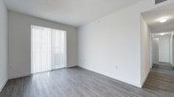 This single-floor space presents a fresh, minimalist aesthetic with a clean and functional design throughout. The main living area is immediately welcoming, featuring smooth grey laminate flooring that lends a contemporary feel underfoot. Crisp white walls provide a neutral, versatile backdrop that complements any style of furnishing or décor. A textured ceiling adds subtle character to the room overhead.

A prominent sliding glass door fitted with full-length vertical blinds serves as a key architectural feature, allowing natural light to filter softly into the space while also offering privacy and light control as needed. The door provides direct access to the outdoors, extending the sense of openness beyond the interior.

The layout is straightforward and efficient, making excellent use of the available footprint on its single level. The overall ambiance is bright and airy, enhanced by the light-toned flooring and white walls that work together to reflect available light and create a sense of spaciousness. The space is move-in ready, offering a clean slate well-suited for a variety of uses and personal styles. Its simple, uncluttered design allows occupants to easily envision and personalize the interior to their preferences.