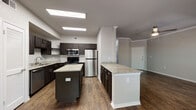 This single-floor residence offers a thoughtfully designed layout with a blend of modern finishes and comfortable living spaces. The expansive kitchen serves as a centerpiece, showcasing dark espresso cabinetry, light granite countertops, a full suite of stainless steel appliances including a gas range, microwave, refrigerator, and dishwasher, along with a central island and a peninsula — providing generous prep and storage space. Hardwood-style flooring extends throughout the main living areas, complementing the neutral wall tones and crown molding details.

The open living and dining area is filled with natural light through large windows, including an elegant arched window, and features a ceiling fan and direct access to an exterior door. The bedroom continues the bright, airy feel with an arched window, ceiling fan, crown molding, and plush carpeting underfoot.

The bathroom is well-equipped with a dual-sink granite vanity, dark cabinetry, a large mirror, and a shower-tub combination. A walk-in closet with wire shelving provides ample storage. Completing the unit is a dedicated laundry area with a full-size washer and dryer, adding everyday convenience to this polished and functional home.