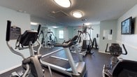This fitness center offers a practical and well-organized space designed to support a full range of workout routines. The room features a dark rubber flooring that provides both durability and safety underfoot, while light-toned walls contribute to a clean and open atmosphere. Large floor-to-ceiling mirrors line one wall, visually expanding the space and allowing users to monitor their form during exercise. The ceiling features a textured finish with flush-mounted lighting that ensures the room is evenly and brightly lit throughout. Cardio equipment, including treadmills and an elliptical machine, is positioned toward the front of the space, offering an accessible starting point for any workout. Toward the rear, a multi-station cable and weight machine provides a comprehensive option for full-body strength training, while a dumbbell rack along the wall supplies a range of free weights for targeted exercises. Natural light filters in through windows, complementing the overhead lighting to create an energizing environment. The space also includes a clearly marked emergency exit, reflecting attention to safety. Overall, this fitness center is thoughtfully arranged to maximize usability and comfort for a variety of fitness levels and training styles.