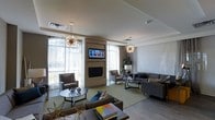 This single-floor space is designed with a modern, upscale aesthetic that combines comfort and sophistication. The open lounge area is anchored by a sleek, built-in fireplace with a wall-mounted flat-screen television above, serving as a natural focal point for the room. Two eye-catching sputnik-style chandeliers hang from the smooth ceiling, casting a warm, ambient glow that complements the generous natural light flowing in through large floor-to-ceiling windows dressed with sheer curtains. The windows frame views of lush outdoor greenery, reinforcing a connection to the surrounding environment. The seating arrangement is thoughtfully curated, featuring a mix of plush sofas and accent chairs in rich brown leather and cool gray upholstery, accented with blue and neutral-toned throw pillows. A light green area rug defines the main seating zone, adding a pop of color against the warm wood-toned flooring. Decorative elements such as sculptural table accents and a potted plant introduce organic texture throughout the space. The overall ambiance is polished and welcoming, making this a functional yet visually refined communal or gathering area.