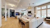 Located at 5948 Aretha Place in Virginia Beach, VA, this thoughtfully designed single-floor space offers a generous, open layout that flows seamlessly between distinct functional zones. The main lounge area is an immediate focal point, anchored by a dramatic floor-to-ceiling stacked stone fireplace wall with a built-in electric fireplace and wall-mounted TV. Light wood flooring runs throughout, complementing the warm, neutral palette. Coffered ceiling details and woven pendant light fixtures add architectural interest overhead, while expansive windows dressed in sheer curtains flood the space with natural light. A substantial rustic wood communal table bridges the lounge and a darker, intimate media nook featuring wall sconces and a technology display panel. The kitchenette is sleek and functional, showcasing light wood cabinetry, crisp white quartz countertops, a gray tile backsplash, brass fixtures, and under-counter refrigeration. A separate seating nook, painted in a rich teal hue, features soaring windows on multiple sides, built-in banquette seating with bold plaid upholstery, and bistro-style tables, creating a bright and inviting social corner. Large black-framed glass doors and transom windows define the entry, reinforcing the space's modern, refined aesthetic throughout.