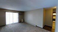 This single-floor space offers a straightforward and functional layout designed for comfortable everyday living. The main living area is finished with wall-to-wall carpet, providing a warm and soft underfoot feel throughout. A large sliding glass door floods the room with natural light and is complemented by full-length vertical blinds, offering both privacy and light control. A wall-mounted air conditioning unit sits above the sliding door, ensuring year-round climate comfort. Along the base of the main wall, a baseboard heating system provides consistent warmth during cooler months, making the space well-equipped for varying seasons. The walls are finished in a neutral beige tone, creating a clean and versatile backdrop that suits a variety of décor styles. A textured popcorn ceiling adds a classic touch overhead. An adjacent area, visible through an opening on one side, hints at additional rooms beyond the main living space. The overall ambiance is calm and neutral, offering a blank canvas ready for personalization. The space is practical and unpretentious, with essential climate control features already in place.