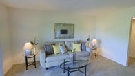 This single-floor residence offers a thoughtfully arranged living space that balances comfort and functionality. The living room serves as the focal point of the home, featuring plush wall-to-wall carpeting that adds warmth underfoot and contributes to the room's cozy ambiance. Neutral, light-toned walls provide a clean and versatile backdrop, allowing the furnishings and décor to stand out naturally. A generously sized sofa anchors the seating area, accented with a mix of patterned and solid throw pillows in navy, yellow, and grey tones that introduce subtle pops of color. A sleek glass-topped coffee table with a metal frame sits at the center, offering both style and practicality. Flanking the sofa are side tables topped with table lamps that provide soft, ambient lighting throughout the room. A decorative framed mirror adorns the wall above the sofa, enhancing the sense of depth and brightness in the space. Tasteful greenery and floral accents are placed throughout, lending a fresh, lively touch to the interior. The overall atmosphere is calm, inviting, and well-balanced, making this space well-suited for relaxing or entertaining.