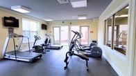 This fitness center offers a clean, functional environment designed to support a variety of cardio-focused workouts. The space is outfitted with quality Precor equipment, including elliptical trainers, a treadmill, and a stationary bike, providing users with a well-rounded selection of cardio machines. The room is finished with durable dark rubberized flooring, ideal for a high-use exercise environment, and features soft yellow walls that contribute to a bright, welcoming atmosphere. Natural light flows in generously through multiple windows and a set of large glass double doors, which also provide direct access to an outdoor area. Recessed panel lighting in the ceiling ensures the space remains well-lit at all times of day. A wall-mounted flat-screen television is positioned for convenient viewing during workouts. The layout is thoughtfully organized to allow comfortable spacing between equipment, enabling users to move freely throughout the room. Emergency exit signage and a security camera reflect a commitment to safety and building standards. Overall, this fitness center is a practical and inviting space suited for daily exercise routines.