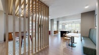 This thoughtfully designed single-floor amenity space blends contemporary aesthetics with functional versatility. The open layout is anchored by a striking full kitchen area featuring a large dark-base island with a white quartz countertop, pendant lighting, and a full run of dark cabinetry complemented by white subway tile. Adjacent to the kitchen, a bright and airy lounge area offers comfortable seating, a mounted flat-screen display, and direct access to an outdoor courtyard through expansive floor-to-ceiling glass doors that flood the space with natural light. A slatted wood room divider adds warmth and visual interest while defining a semi-private dining nook, which features a bold green accent wall, pendant globe lighting, and a wood-slat ceiling detail. A cozy banquette dining area with teal upholstered seating, a white tabletop, and expressive botanical wallpaper sits beside large windows offering urban views. The space transitions seamlessly between zones via light wood plank flooring throughout. Thoughtful design details—including curated artwork, lush indoor plants, and a mix of mid-century modern furnishings—create a welcoming and dynamic communal environment suited for socializing, dining, or casual gatherings.
