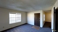This single-floor space presents a straightforward and functional layout centered around a spacious main living area. The room is finished with wall-to-wall blue-gray carpeting that extends throughout, while a small section of hard surface flooring marks the entryway just inside the dark, six-panel front door — a classic design detail that adds a traditional character to the space.

The textured popcorn ceiling runs across the entire room, a common feature that adds dimension overhead. Two windows are positioned along the walls, allowing natural light to filter in and brighten the interior. The neutral, light-colored walls provide a clean and versatile backdrop suitable for a variety of furnishing styles.

The entry area is well-defined by the transition in flooring, creating a natural boundary between the front door and the main living space. The open layout offers flexibility in how the space can be arranged and utilized. Overall, the space is practical and move-in ready, with a simple, no-frills aesthetic that prioritizes function and comfort.