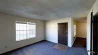This single-floor space presents a straightforward and functional layout centered around a spacious main living area. The room is finished with wall-to-wall blue-gray carpeting that extends throughout, while a small section of hard surface flooring marks the entryway just inside the dark, six-panel front door — a classic design detail that adds a traditional character to the space.

The textured popcorn ceiling runs across the entire room, a common feature that adds dimension overhead. Two windows are positioned along the walls, allowing natural light to filter in and brighten the interior. The neutral, light-colored walls provide a clean and versatile backdrop suitable for a variety of furnishing styles.

The entry area is well-defined by the transition in flooring, creating a natural boundary between the front door and the main living space. The open layout offers flexibility in how the space can be arranged and utilized. Overall, the space is practical and move-in ready, with a simple, no-frills aesthetic that prioritizes function and comfort.