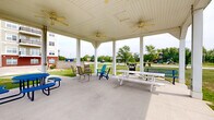 Located at 1627 Cottleville Pkwy in Cottleville, Missouri, this outdoor community space offers a well-appointed gathering area designed for relaxation and socializing. At its centerpiece is a spacious covered pavilion supported by white columns and outfitted with multiple ceiling fans, providing comfortable shade and airflow throughout the warmer months. The pavilion accommodates a variety of seating arrangements, including picnic tables, Adirondack chairs, and casual lounge seating, making it suitable for both small gatherings and larger group events. A gas grill stationed nearby adds to the pavilion's utility as an outdoor entertainment hub. Just beyond the covered structure, a brick-edged fire pit is encircled by vibrant teal Adirondack chairs, creating an inviting focal point for evening gatherings. The surrounding grounds feature expansive, well-maintained lawns dotted with young trees and manicured shrubs, offering an open and airy feel with views stretching toward a playground and additional green space in the distance. A multi-story residential building is visible in the background, contextualizing the space within a broader community setting. The combination of functional amenities and thoughtfully landscaped outdoor areas makes this a versatile and welcoming space for residents and visitors alike.