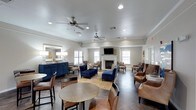 This single-floor leasing center offers a thoughtfully designed and inviting environment for prospective residents and visitors alike. The main lounge area serves as the heart of the space, featuring a classic white fireplace flanked by windows that allow natural light to fill the room, creating a warm and welcoming ambiance throughout.

The seating arrangement is both varied and stylish, combining plush navy blue armchairs and a coordinating sofa, rich brown leather accent chairs, and patterned upholstered seating to accommodate different preferences and group sizes. A natural fiber area rug anchors the central seating area, while round bistro-style tables near the entrance provide a casual spot for conversation or paperwork.

A bold navy blue credenza along one wall adds a striking design element, complemented by decorative mirrors and accent lighting that enhance the room's refined aesthetic. Crown molding trims the walls, contributing to the polished, upscale feel of the interior. Dual ceiling fans with integrated lighting ensure comfort across the generously sized room, while recessed lighting throughout provides consistent illumination. Large windows on multiple walls invite views of the surrounding exterior and reinforce the open, airy character of this well-appointed leasing center.