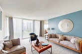 This well-appointed two-bedroom, two-bathroom residence in Silver Spring, Maryland offers a thoughtfully designed single-floor layout. The expansive living room is a standout feature, showcasing floor-to-ceiling windows and sliding glass door access to a balcony, flooding the space with natural light and providing panoramic views of the surrounding cityscape. A striking blue accent wall adds a bold design element to the living area, which flows seamlessly into the dining room. The dining room benefits from its own large window with city views and is highlighted by an elegant chandelier overhead. The kitchen is outfitted with white cabinetry, dark countertops, a full suite of appliances, and tile flooring, offering a clean and functional workspace. The generously sized primary bedroom features plush carpeting, warm neutral tones, and a large window. A second bedroom provides additional flexibility. Two full bathrooms, three closets, and an in-unit laundry room round out the layout, providing excellent storage and everyday convenience. Throughout the residence, neutral tones, carpet flooring in the living areas, and abundant natural light contribute to a comfortable and inviting atmosphere.