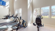 This fitness center is a thoughtfully designed, single-floor space that combines an open, airy layout with a comprehensive selection of professional-grade equipment. The room's most striking architectural feature is its soaring ceiling height, which lends a spacious, energizing atmosphere enhanced by an abundance of natural light streaming through multiple large windows. Views of the surrounding exterior add a welcoming sense of connection to the outdoors.

The flooring throughout is a durable speckled composite, well-suited for high-traffic fitness use. A full-length wall mirror along one side visually expands the space while allowing users to monitor their form during exercise. Colorful framed artwork displayed near the upper wall adds a vibrant, motivating touch to the otherwise clean, neutral palette of white and light beige walls.

The cardio zone is well-appointed with multiple True-brand treadmills and stationary bikes, including both upright and recumbent options, each equipped with individual display screens. A dedicated rowing machine rounds out the aerobic offerings. The strength training area features a tiered dumbbell rack with a full range of free weights, alongside several selectorized machines including leg extension, cable, and back extension units. Wall-mounted flat-screen televisions are positioned throughout for entertainment. The space also includes restroom access and a clearly marked exit.