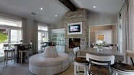 Located at 2155 Benton Blvd in Savannah, Georgia, this well-appointed single-floor space offers a refined and inviting atmosphere defined by thoughtful architectural details and high-quality finishes. The centerpiece of the main living area is a dramatic floor-to-ceiling stacked-stone fireplace, which anchors the room and serves as a striking focal point. A wall-mounted television is seamlessly integrated into the stonework above the firebox, blending functionality with design. Soaring vaulted ceilings accented by exposed dark wood beams lend the space a sense of grandeur and warmth. Built-in shelving and cabinetry with louvered lower panels provide both display space and practical storage, adding a classic touch to the décor. The open-concept layout flows naturally from the lounge area — featuring a distinctive circular upholstered ottoman — into a dining area furnished with a round table and mixed seating, creating a comfortable and social environment. Large windows dressed with neutral drapery allow natural light to filter in, enhancing the bright and airy feel of the interior. Recessed lighting throughout further illuminates the space, while a neutral color palette of warm grays and whites ties all design elements cohesively together.