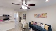 This 1-bedroom, 1-bathroom residence offers a thoughtfully arranged single-level layout that maximizes every square foot. The open living area serves as the heart of the home, featuring light-toned hardwood-style flooring, neutral walls, and a ceiling fan with integrated lighting that keeps the space bright and comfortable. Colorful wall art and carefully chosen furnishings add warmth and personality to the room. Recessed lighting throughout enhances the clean, modern aesthetic. The living space flows naturally toward the kitchen, which is partially visible and features light cabinetry, contributing to the home's cohesive, airy feel. A dedicated workspace with a bar-height desk and stool provides a functional area for productivity. The bedroom, visible in the background, continues the consistent flooring and neutral palette, creating a calm and restful retreat. The overall design balances practicality with style, making efficient use of the available space while maintaining a welcoming ambiance. This unit is well-suited for those seeking a comfortable, move-in-ready home with modern touches in a compact and efficient footprint.