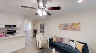 This 1-bedroom, 1-bathroom residence offers a thoughtfully arranged single-level layout that maximizes every square foot. The open living area serves as the heart of the home, featuring light-toned hardwood-style flooring, neutral walls, and a ceiling fan with integrated lighting that keeps the space bright and comfortable. Colorful wall art and carefully chosen furnishings add warmth and personality to the room. Recessed lighting throughout enhances the clean, modern aesthetic. The living space flows naturally toward the kitchen, which is partially visible and features light cabinetry, contributing to the home's cohesive, airy feel. A dedicated workspace with a bar-height desk and stool provides a functional area for productivity. The bedroom, visible in the background, continues the consistent flooring and neutral palette, creating a calm and restful retreat. The overall design balances practicality with style, making efficient use of the available space while maintaining a welcoming ambiance. This unit is well-suited for those seeking a comfortable, move-in-ready home with modern touches in a compact and efficient footprint.