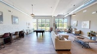 Located at 409 East Butler Avenue in New Britain, PA, this single-floor community living room is a thoughtfully designed gathering space that blends modern aesthetics with a warm, inviting atmosphere. The open-plan layout is anchored by rich hardwood flooring that runs throughout, complementing the neutral warm-toned walls and high ceilings that lend the space a generous sense of volume.

Large, floor-to-ceiling windows with black steel framing line one side of the room, flooding the interior with natural light and offering unobstructed views of the surrounding greenery. A full-size pool table serves as a central recreational feature, positioned near the sun-drenched window wall alongside a seating cluster of accent chairs ideal for casual conversation.

A long bar-height counter with leather-upholstered stools runs along a stone-accented accent wall, providing a dedicated workspace or social perch. Framed artwork and decorative touches, including equestrian-themed paintings and local photography, add character and personality throughout.

The lounge area features a generous sectional sofa and coffee table arranged over a woven area rug, creating a comfortable retreat for relaxation. A wall-mounted television, media console, and a dining table with chairs round out the amenities. Pendant lighting fixtures with an industrial character hang from the ceiling, completing the space's contemporary yet approachable design.