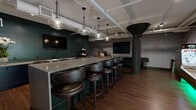 Located at 40 Locke St in Haverhill, Massachusetts, this thoughtfully designed entertainment space offers a dynamic blend of social and recreational amenities across a single expansive floor. The interior showcases an industrial-chic aesthetic, with exposed ceiling ductwork and piping contrasted against warm hardwood flooring and richly patterned wallcoverings that lend texture and character throughout.

A central bar area anchors the space, featuring a long concrete-topped counter lined with leather bar stools and illuminated by globe pendant lights, with deep green tile accents and wall-mounted televisions creating an inviting lounge atmosphere. Distinctive funnel-shaped structural columns serve as bold architectural focal points, adding an industrial artfulness to the design.

The game zone includes two billiards tables, a ping-pong table, and a skee-ball lane with LED accent lighting, providing ample entertainment options for groups. A comfortable lounge area is furnished with tufted leather sofas, area rugs, and a statement sputnik-style chandelier, centered around a mounted TV wall panel finished in geometric tiled detailing.

Large industrial-style windows allow natural light to filter into the lounge section, while potted tropical plants add organic warmth to the space. The overall ambiance is sophisticated yet playful, making this a well-appointed venue suited for social gatherings and recreational use.