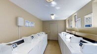This single-floor communal laundry facility is designed with practicality and efficiency in mind. The room features two parallel rows of modern, white top-loading washing machines and dryers neatly arranged along both walls, providing ample capacity for residents. The space is finished with neutral beige walls and a smooth, easy-to-clean floor, contributing to a clean and utilitarian aesthetic. Overhead fluorescent lighting ensures the room is well-lit throughout, creating a bright and functional environment. A window along one wall allows for a degree of natural light to supplement the artificial lighting. Instructional guides are posted on the wall near the machines for user reference, and a utility sink is accessible near the window side. A centrally located door at the far end of the room provides additional access. The overall layout is thoughtfully organized to allow users to move freely between machines, making the laundry process straightforward and convenient. This facility reflects a commitment to providing residents with a well-maintained, fully equipped shared amenity space.
