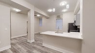 This single-floor unit offers a thoughtfully designed open-concept layout that efficiently combines the kitchen and living area into one cohesive space. Light gray hardwood-style flooring runs throughout, creating a clean, contemporary foundation that unifies the rooms.

The kitchen is a standout feature, equipped with stainless steel appliances including a refrigerator, range, and microwave. Gray shaker-style cabinetry provides ample storage, complemented by white countertops and a peninsula with an integrated sink, offering both additional workspace and a natural division between the kitchen and living areas. Decorative pendant lighting hangs above the peninsula, adding a refined touch, while recessed ceiling fixtures provide consistent ambient illumination throughout the open space. Windows fitted with horizontal blinds allow natural light to filter in.

A structural column near the center of the main living area adds an architectural element to the otherwise open floor plan. The living room provides a versatile, open area adjacent to the kitchen.

The bathroom maintains the unit's neutral palette with a white vanity, a mirrored medicine cabinet, and matching hardwood-style tile flooring, offering a clean and functional design.