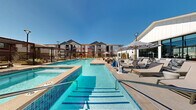 Located at 10670 Jake Jabs Blvd in Firestone, Colorado, this resort-style pool amenity area is a standout outdoor destination designed for relaxation and recreation. The centerpiece is a large, crystal-clear swimming pool complemented by a separate spa, both finished with striking blue tile and surrounded by a spacious concrete sundeck. The deck is generously appointed with lounge chairs, Adirondack-style seating, and umbrella-shaded areas, providing ample space for sunbathing and socializing under Colorado's open blue skies.

Adjacent to the pool, a well-appointed outdoor lounge patio features comfortable upholstered seating arranged around a coffee table, set against the backdrop of a modern farmhouse-style clubhouse building with large floor-to-ceiling windows and stone accent details. Pergola structures and steel privacy screens offer both shade and architectural interest throughout the space.

A dedicated recreation lawn area provides room for lawn games, including cornhole, and is lined with tall light poles for evening use. Landscaped pathways with native plantings, ornamental grasses, and decorative rock borders weave through the community, connecting the amenity spaces. The overall design reflects a cohesive, contemporary aesthetic that blends modern farmhouse architecture with functional, resort-caliber outdoor living.