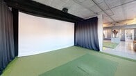 Located at 127 N 17th St in Richmond, Virginia, Baker's 19th Hole is a single-floor golf entertainment space that blends industrial architecture with bold, modern design. The interior is defined by exposed concrete ceilings and structural elements, polished concrete floors, and striking navy blue geometric-patterned accent walls trimmed in gold — creating a sophisticated yet relaxed atmosphere throughout.

The main area centers on a multi-hole indoor putting green with red-flagged cups, surrounded by a comfortable lounge zone featuring a leather sofa, bar-height seating, and a decorative "GOLF" wall installation, all bathed in natural light from large street-facing windows. Adjacent to the lounge is a dedicated golf simulator bay, equipped with a full projection screen and overhead launch monitor, turf flooring, and a heavy curtain divider for focused play.

A compact self-serve market near the entrance offers snacks, beverages, and sundries through an automated checkout kiosk, making it easy for guests to grab refreshments. The entryway includes a cushioned bench and a branded "Baker's 19th Hole" wall medallion, welcoming visitors with character. The space also connects visually to an adjacent bright lobby area. Every detail of this venue reflects a cohesive design vision built for golf enthusiasts and casual visitors alike.
