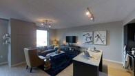 This single-floor space showcases a thoughtfully designed open-concept layout that connects the living area and kitchen into one cohesive environment. The living room is anchored by a plush sofa and a tan leather accent chair arranged around a dark navy shag area rug, creating a cozy yet sophisticated seating area. A wall-mounted flat-screen television sits atop a sleek media console, flanked by decorative accents and table lamps that contribute to the warm ambiance. Two abstract framed artworks hang prominently on the wall, adding a contemporary artistic touch to the neutral grey palette throughout. A sculptural chandelier and adjustable track lighting provide layered illumination across the space. Natural light filters in through a large window, offering views to the exterior. Transitioning toward the kitchen, a dark-finished island with a light granite countertop serves as both a functional workspace and a visual divider between the living and kitchen zones. Light wood-toned flooring runs throughout, complementing the modern, understated aesthetic. The overall atmosphere is polished and inviting, balancing comfort with contemporary design sensibility.